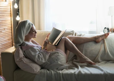Frau sitzt in Bademantel und mit Handtuch auf dem Kopf auf einer Couch und liest ein Buch