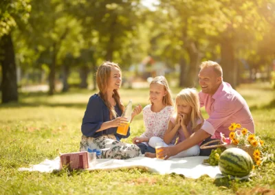 Mann sitzt mit Frau und zwei Mädchen auf einer Picknick Decke in einem Park. Im Hintergrund befinden sich Böume, auf der Decke sind eine Wassermelone, Weintrauben und eine Flasche mit Saft